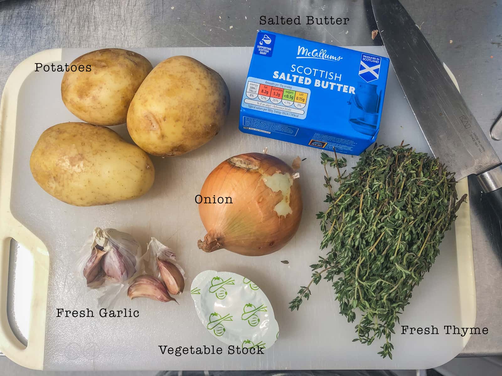 Ingredients on a board to prepare boulangere potatoes consisting of potatoes, garlic, onions, butter, vegetable stock and fresh thyme.