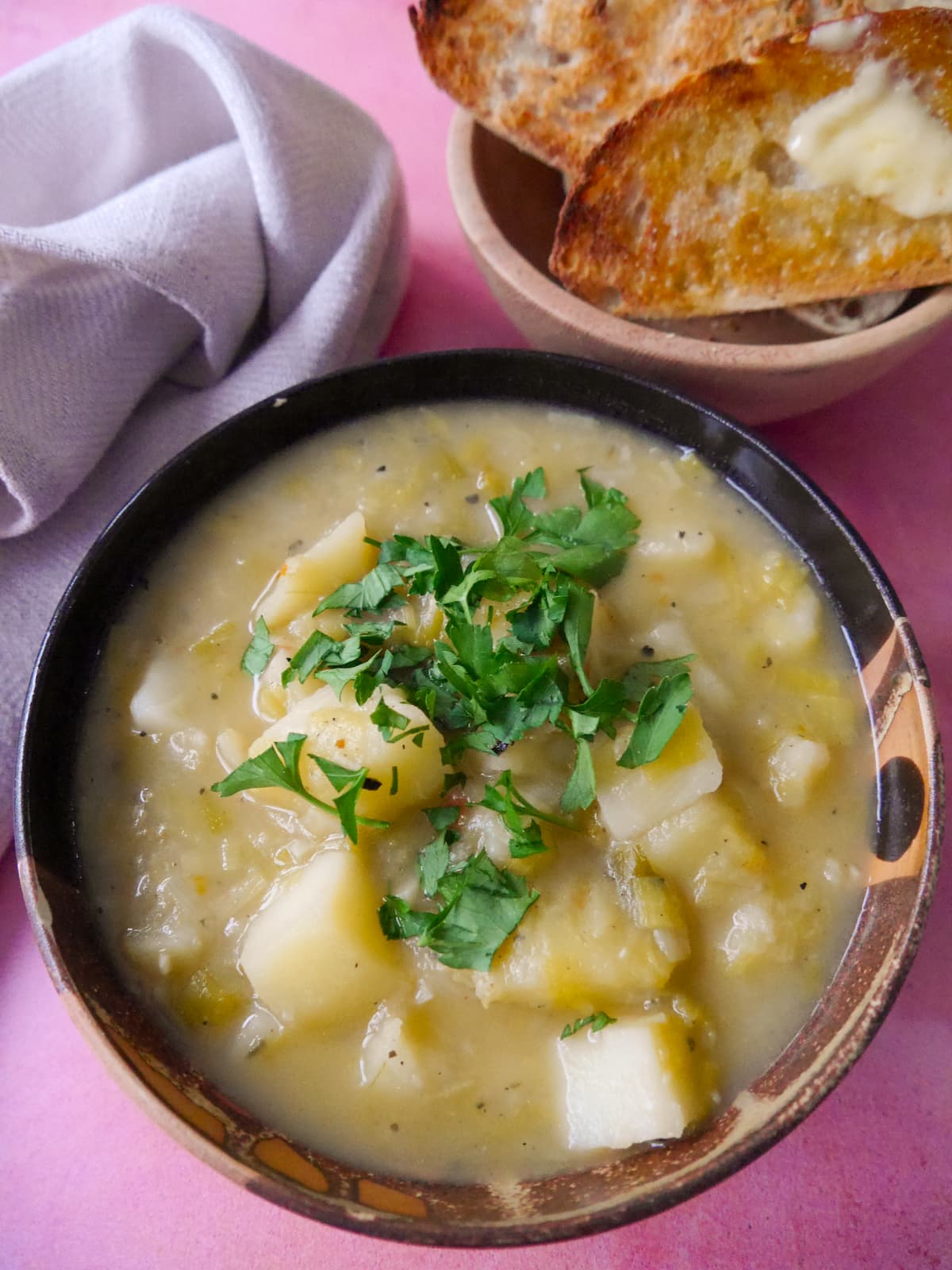 A bowl of rustic leek and potato soup topped with a garnish of chopped parsley, with a side of buttered toast.