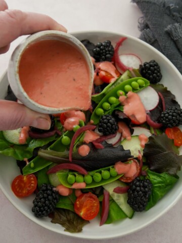 A bowl of mixed salad leaves and vegetables with a small jug of fresh raspberry vinaigrette dressing being poured over.