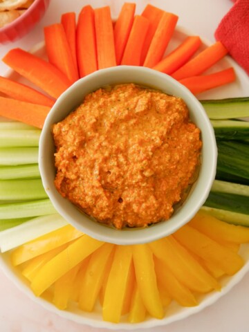 A bowl of roasted red pepper dip served with a variety of fresh vegetable sticks, including bell pepper, cucumber, celery and carrot.